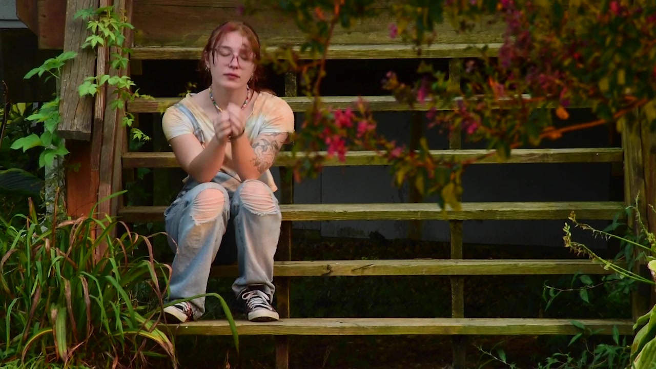 A young woman sits on wooden steps beside a green garden in Kick Back